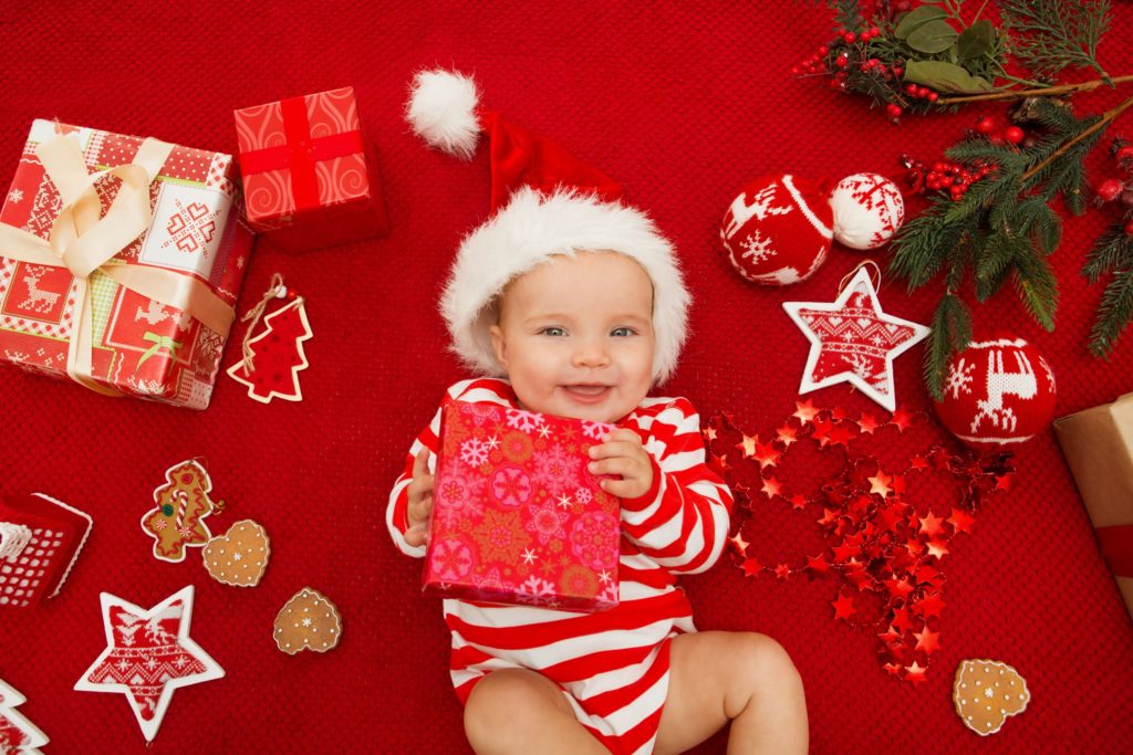 Cute baby smiling in Santa hat and red and white onesie holding a present whilst surrounded by Christmas present and decorations