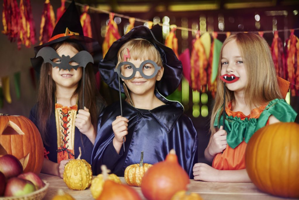 Two girls and one boy dressed up for a Halloween party. They are surrounded by pumpkins and party decorations. All are holding up Halloween eye masks to their face or mouth
