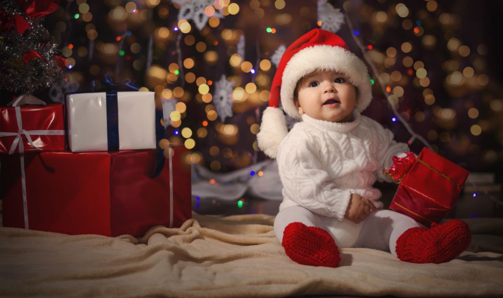 Cute baby sat by Christmas tree holding a present and wearing a Santa hat