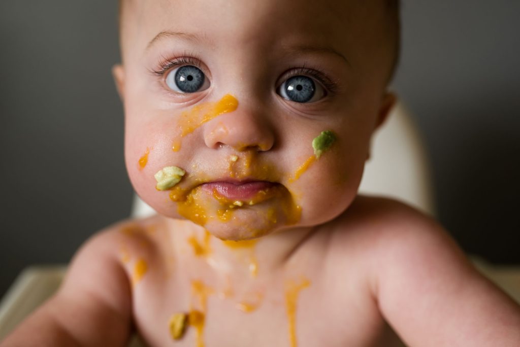Baby with big blue eyes with baby food all over their face
