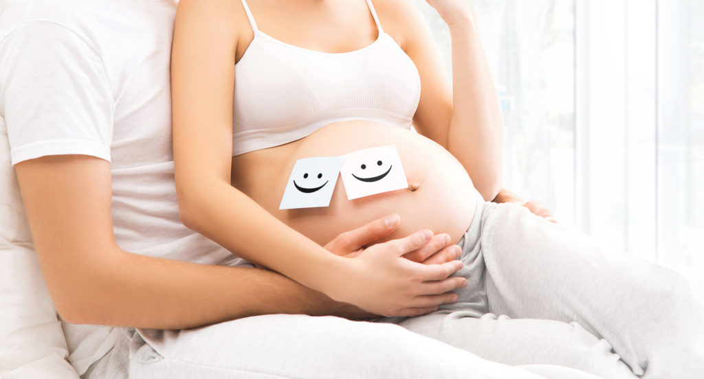 Man lovingly holding his partner's baby bump. The baby bum has two smiley face post it notes stuck to it to resemble having twins