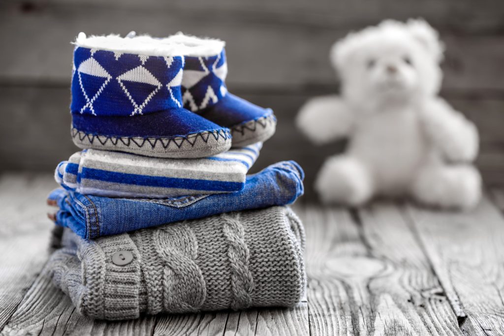 Baby winter clothes like jumper, pants, onesie and booties placed on top of each other whilst a teddy features in the background of the picture