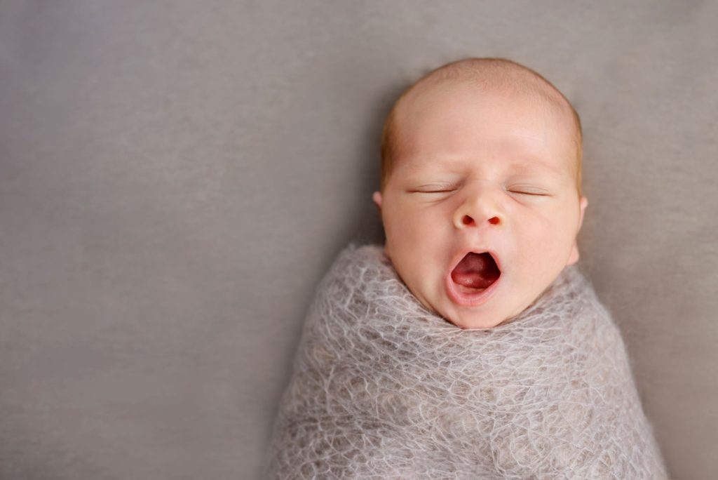 Newborn baby yawning whilst in a grey swaddle