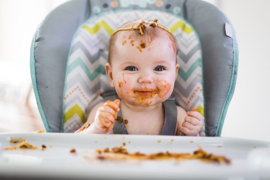 Little girl smiling in highchair with food mess all over her face and highchair table