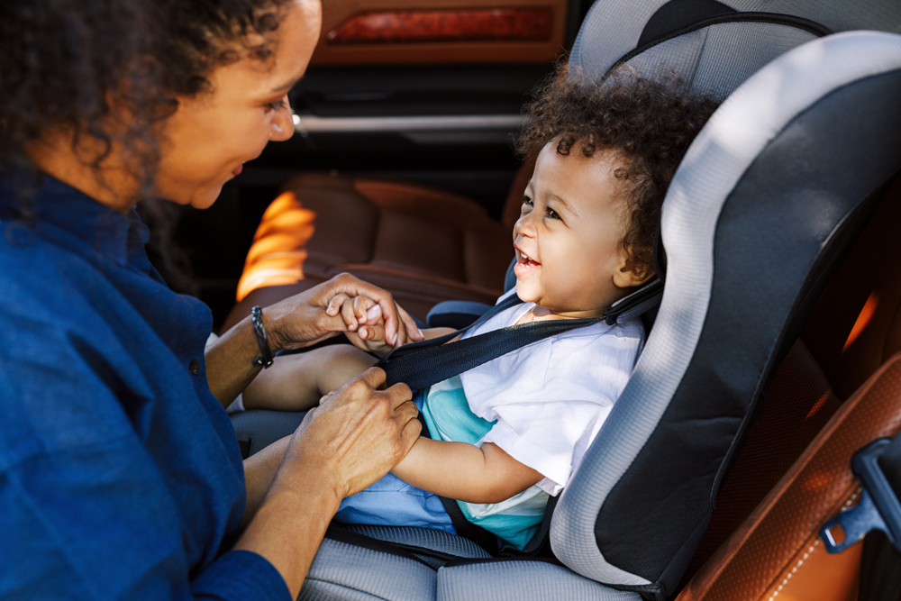 Side View Of A Mother Helping Toddler Get Buckled Into Car Seat
